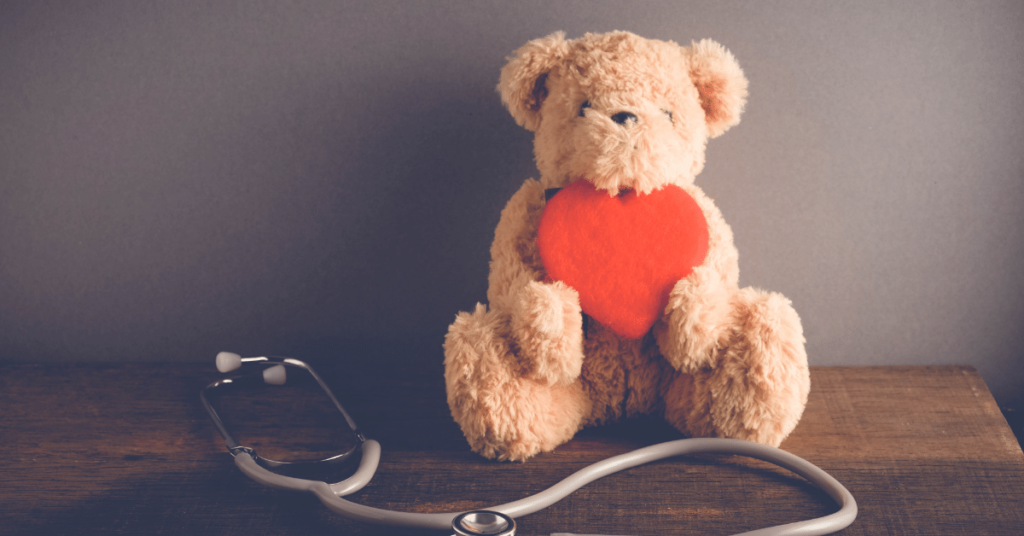 A teddy bear sits on a table holding a large heart. A stethoscope sits in front