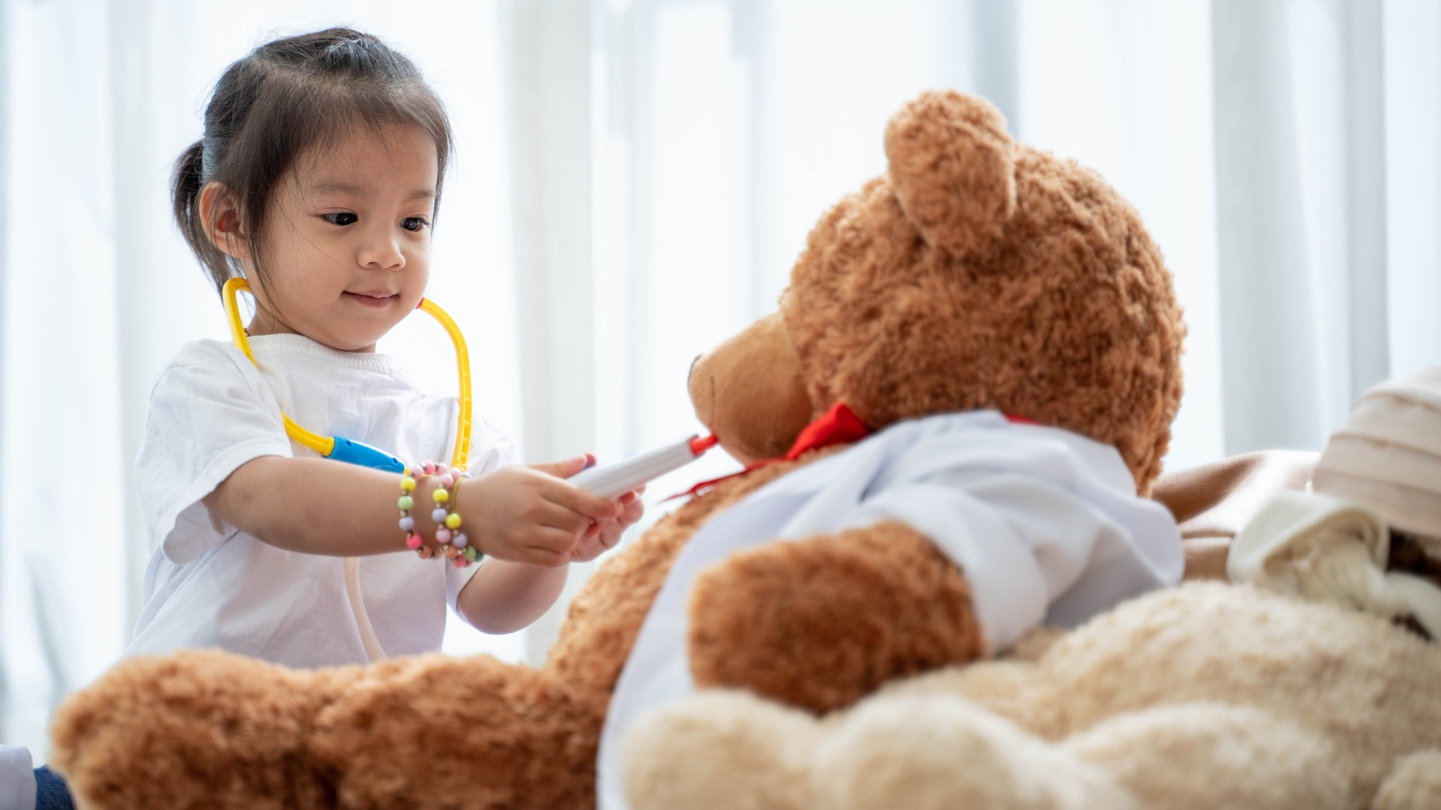 a young child uses a toy stethoscope and thermometer to care for a large teddy bear