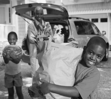 a family of three stand behind their open van in the driveway. a young boy holds a grocery bag full of food, another young child holds a watermelon in the background, while an adult stands further back