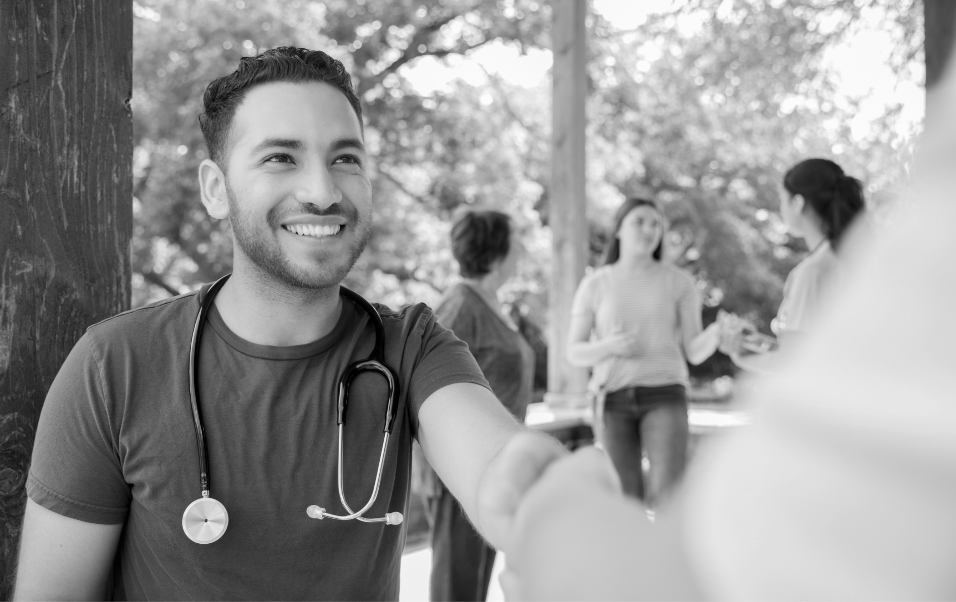 a young adult wearing a stethoscope stands outside speaking to someone. a group chats in the background