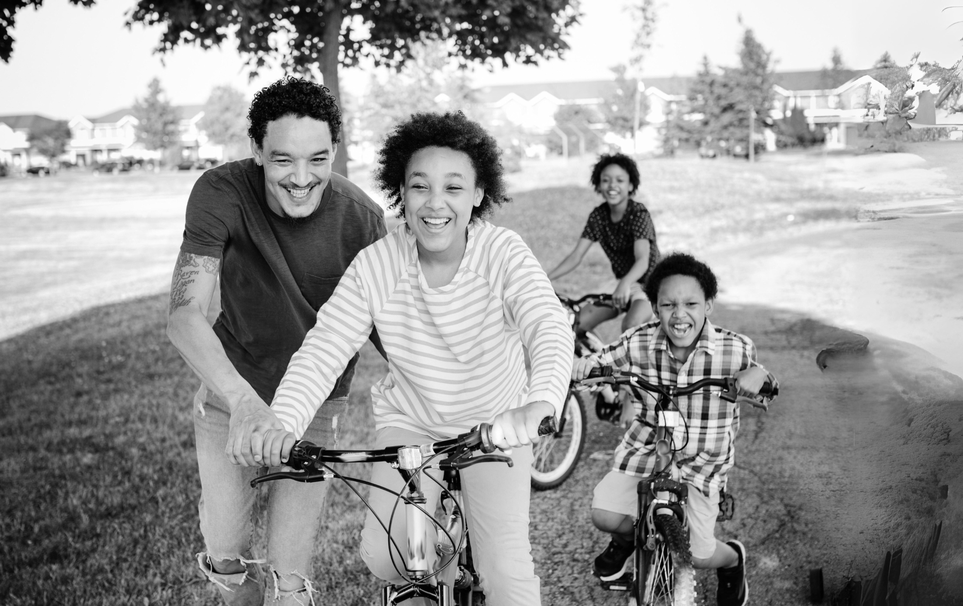 an adult helps a child ride a bike, while 2 other kids ride behind
