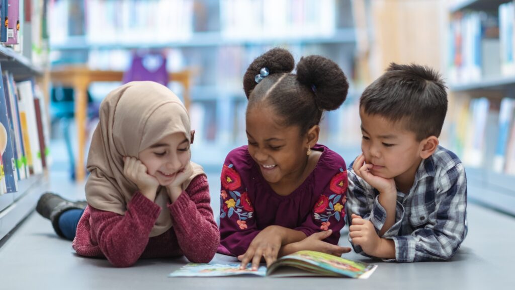 A group of 3 diverse children lay on the floor of a library looking at a book together. a child tax credit in Virginia would help children and families across the commonwealth