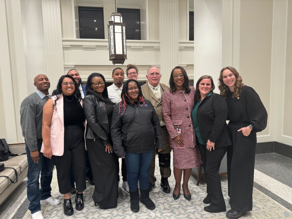 A group photo of fines and fees and decriminalization of poverty advocates, including members from REAL Life, Help Me Help You, Legal Aid Justice Center, advocacy partners, and individuals with lived experience. The group is standing in a well-lit indoor setting with white walls and large windows. Senator Williams Graves is in the center, wearing a pink tweed suit. The group is smiling 