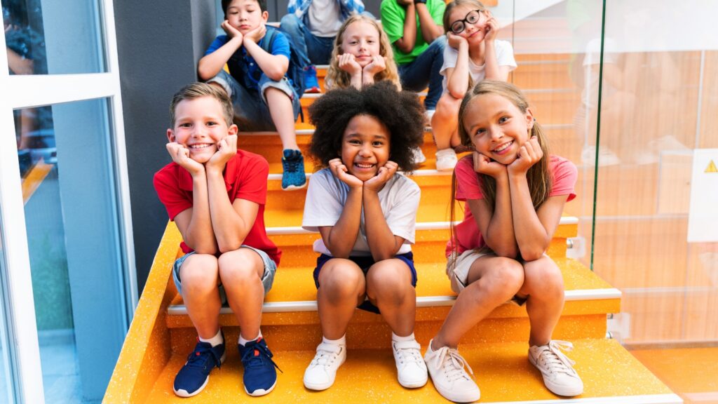 a group of young, diverse students sit on stairs in rows of three. The students wear a variety of facial expressions with those in the front smiling. Two students in the second row have subtle smiles while the 3rd slightly puts with their eyes downcast