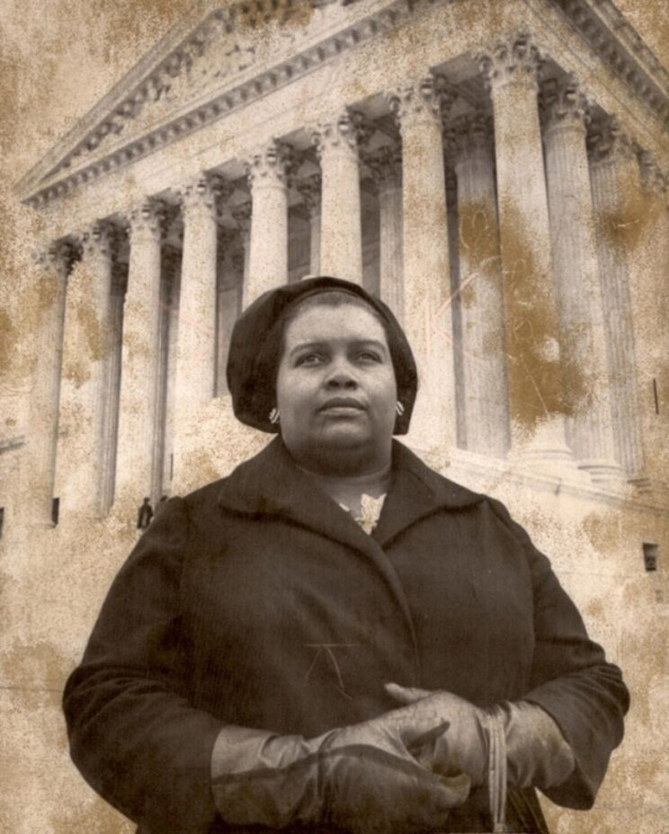 A Black woman wearing a coat, gloves, and a hat stands in front of a building with several large columns