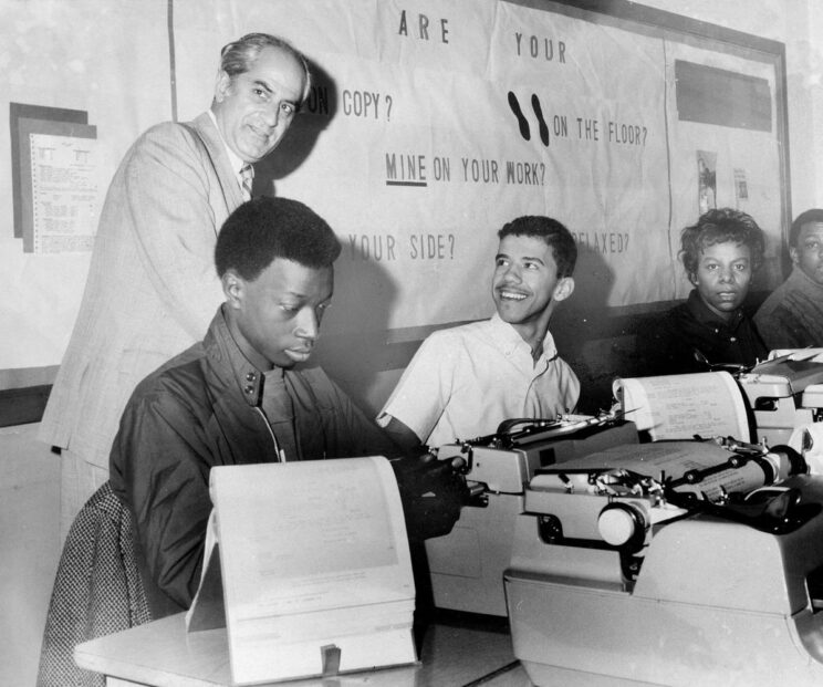 Several people sit at a table in front of word processing machines. A man stands in the back, looking over his shoulder toward the camera, while one of the people sitting look up and smile towards the man.