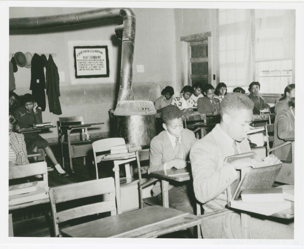 Several rows of desks in a classroom. the vast majority are occupied by students. A round, metal device sits in the center-back of the classroom with a large pipe extending up and across the wall