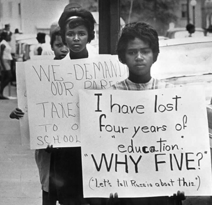 Three Black students are clearly seen, each carrying a sign. The first says 