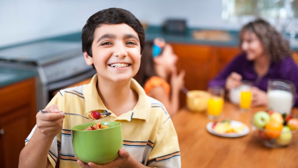 a young boy smiles at the camera while holding a spoon above a bowl with fruit and granola. Another young child and adult are blurred in the background