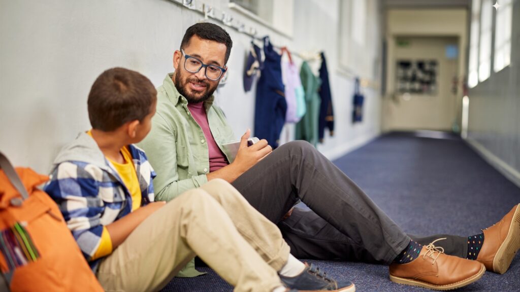 a young child and adult sit in a hallway talking. in the background, jackets hang from a wall