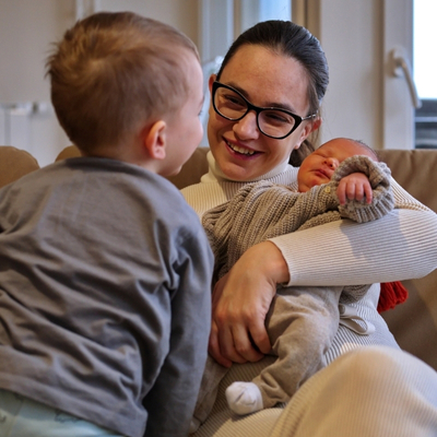 A woman wearing glasses holds an infant while a young toddler sits facing her on a couch. Virginia refundable state tax credits help working families of many sizes make ends meet.