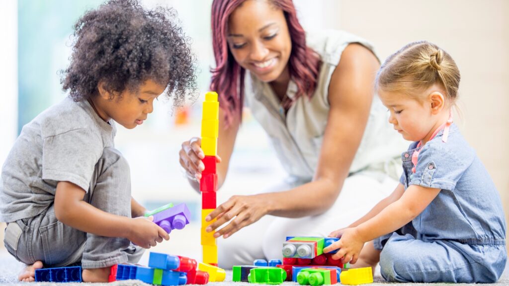 Smiling adult playing with two young children as they build a colorful block tower on the floor. Greater access to high-quality child care in Virginia could improve school readiness and higher high school graduation rates.
