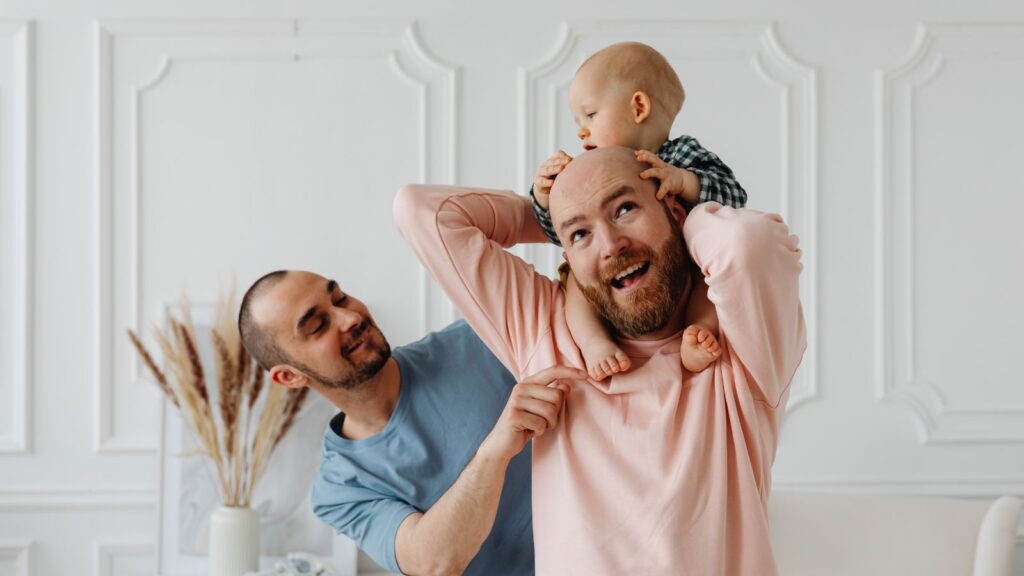 A joyful scene of parents and their baby. One father holds the baby on his shoulders with laughter, while the other playfully interacts. Bright, cozy room setting.