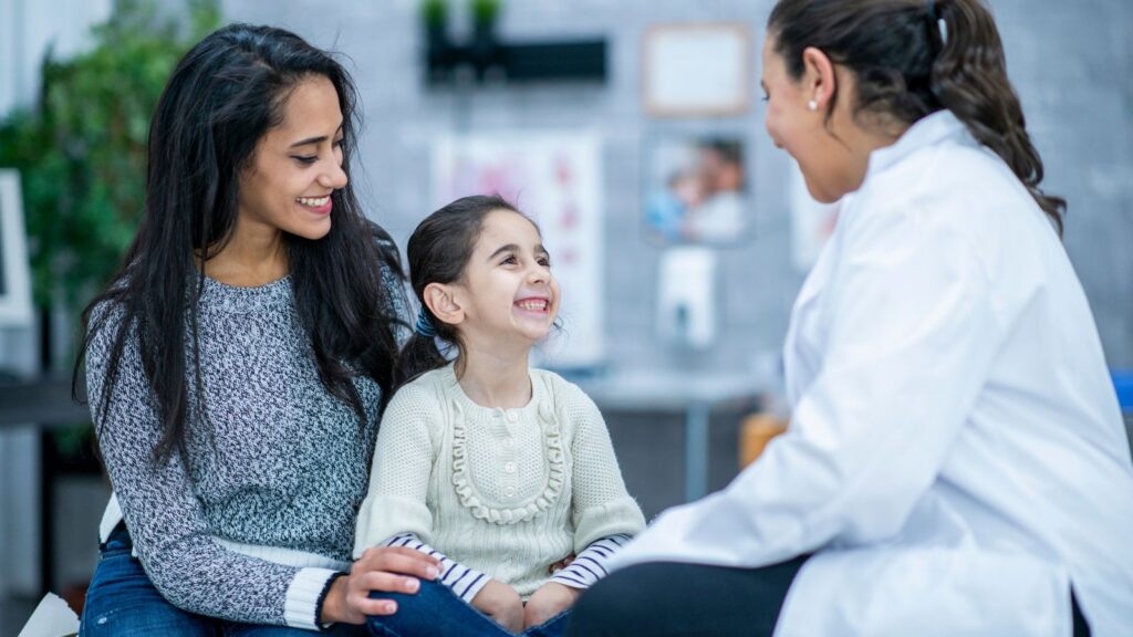 A smiling young girl sits on her mother's lap during a pediatric appointment, both radiating warmth and ease as they engage with a female physician in a white coat. The scene reflects a compassionate, family-centered approach to care — where children feel safe and parents feel supported.