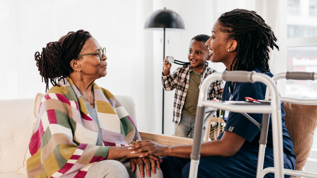 A home health nurse in scrubs sits with an older woman wrapped in a colorful blanket, holding her hands warmly during a home visit, while a young boy playfully holds up a stethoscope in the background. A walker rests nearby. The scene speaks to the full arc of care — from the very young to older adults — and the vital role that health safety nets play in keeping families whole, connected, and cared for in their own homes and communities.