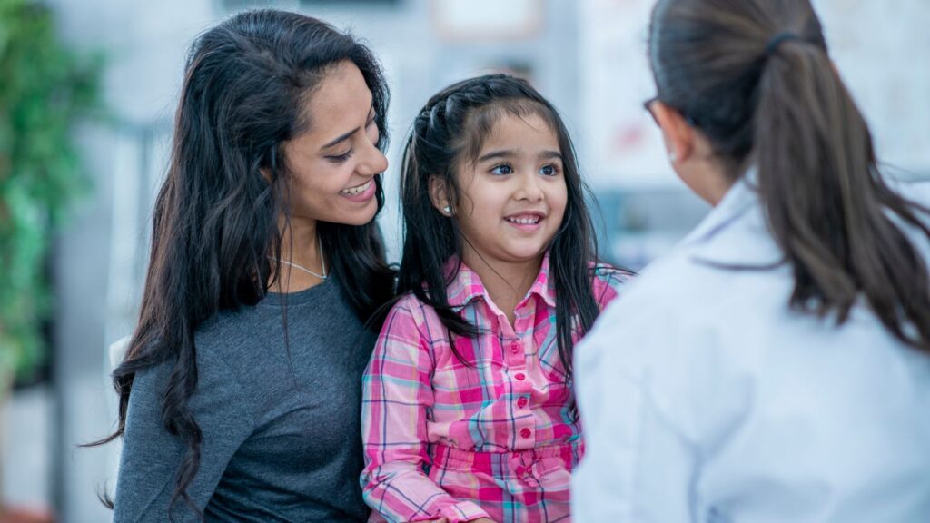 A smiling young girl sits on her mother's lap during a pediatric appointment, both radiating warmth and ease as they engage with a female physician in a white coat. The scene reflects a compassionate, family-centered approach to care — where children feel safe and parents feel supported.