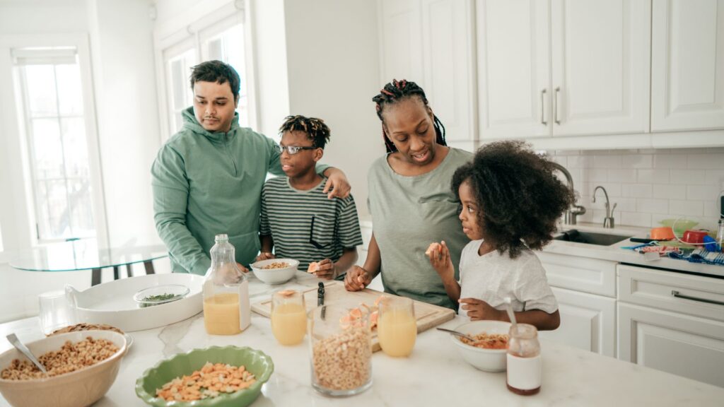 A family gathers around a kitchen counter covered with breakfast foods, including cereal, juice, and fruit — everyday moments shaped by the cost of living."