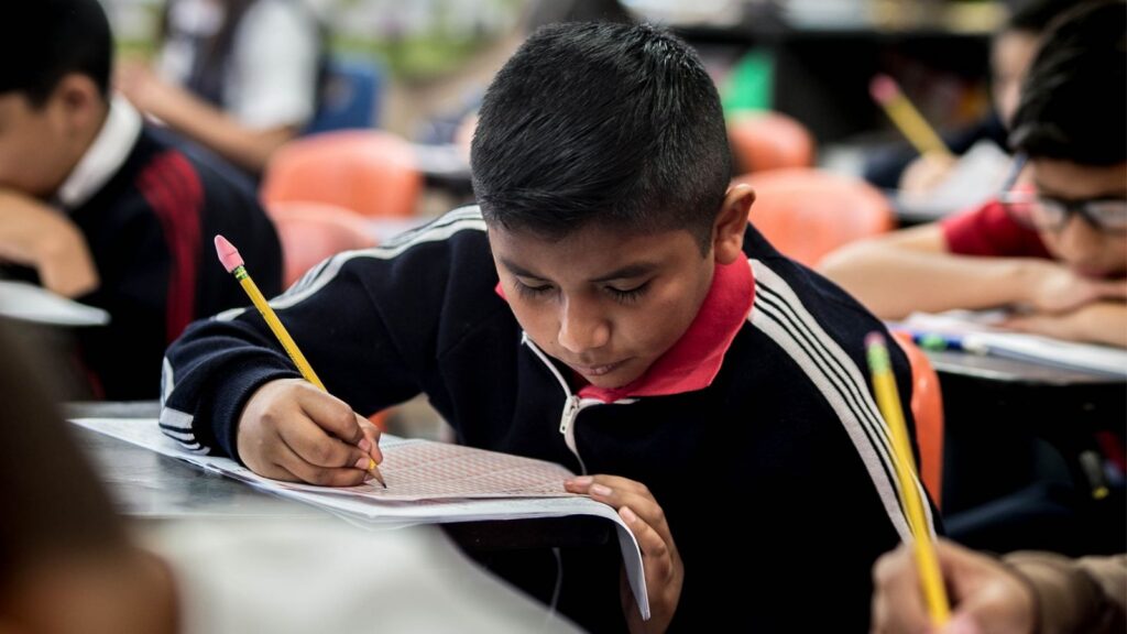 A young student concentrates while filling out a worksheet in a classroom, reflecting the daily learning experiences that adequate school funding helps support.