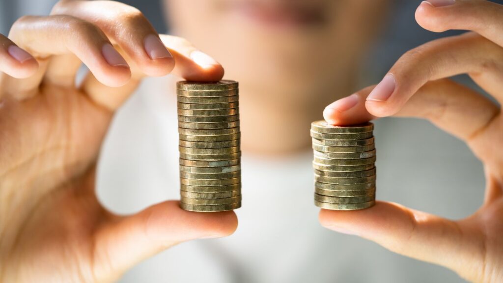 Close-up of two hands holding unequal stacks of gold coins side by side, one noticeably taller than the other.
