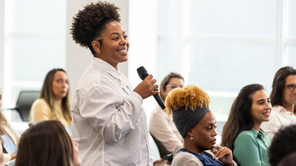 A woman stands and speaks into a microphone at a community meeting, smiling, while attentive audience members sit around her in a bright, window-lit room.