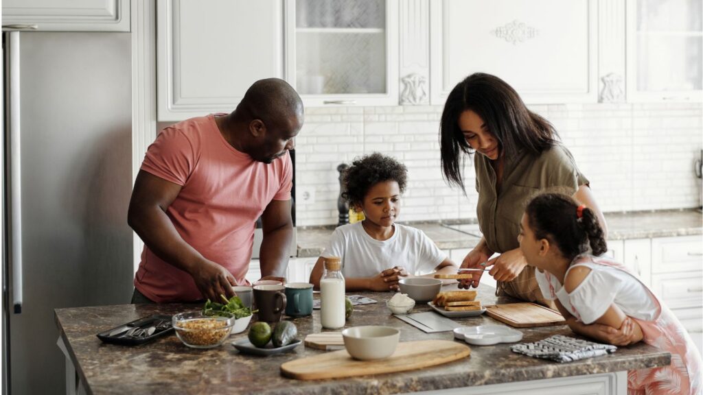A father, mother, and two children gather around a kitchen island preparing breakfast together, with toast, fruit, granola, and milk spread across the counter.