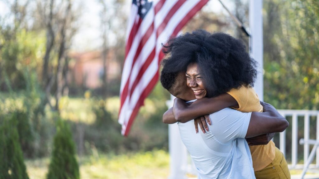 Two people embrace joyfully in a front yard, an American flag waving behind them — a homecoming moment.