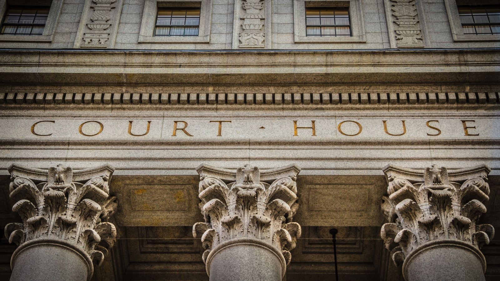 Image of a close up shot of the front of a court house. Tops of the columns can be seen as well as the two words "Court House" in all caps.